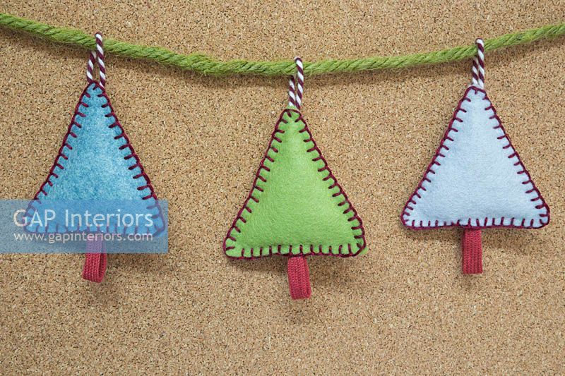 Making stitched felt christmas decorations - miniature christmas trees made from felt and decorative string, hanging against a cork board