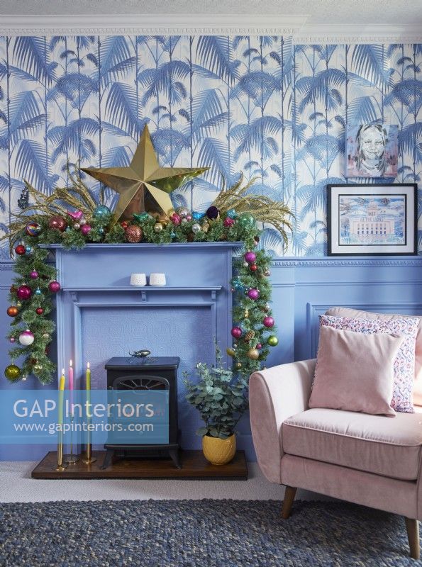 Living room detail showing a mantlepiece decorated for Christmas, jungle print wallpaper and blue panelling.