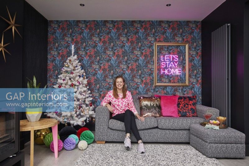 Open plan living room decorated for Christmas. Cosy sofa with colourful cushions, patterned wallpaper, black walls and a wood burner stove.