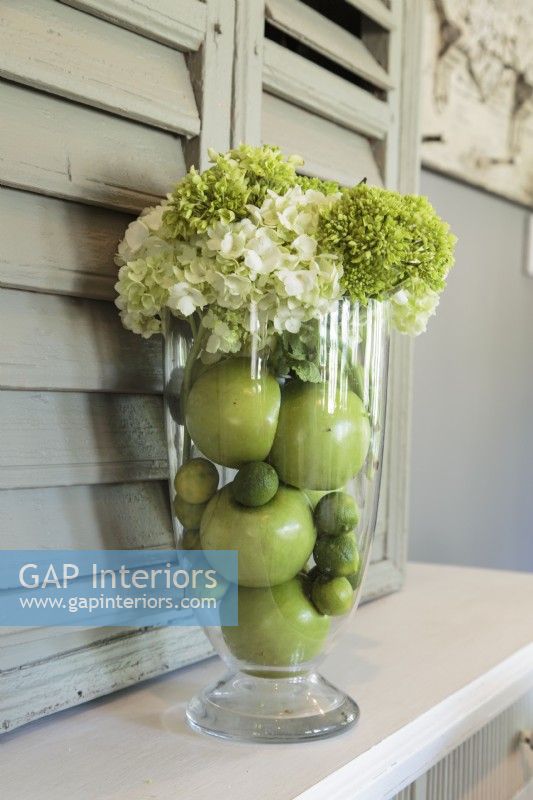 A clear vase holds a simple arrangement of hydrangeas, apples and limes in the same color family that brings the garden in.