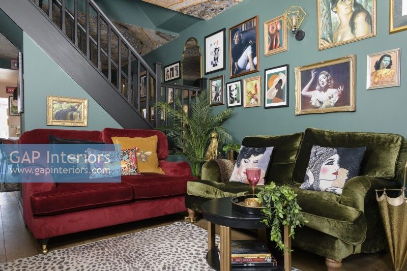 Open plan green living room with red and green velvet sofas in front of an open staircase and a salon style gallery art display wall