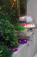 Rendered raised bed beside fireplace with Liriope muscari, candles and a large glass bowl with gerberas in garden at night