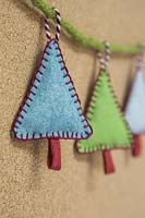 Making stitched felt christmas decorations - miniature christmas trees made from felt and decorative string, hanging against a cork board