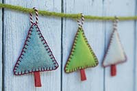 Making stitched felt christmas decorations - miniature christmas trees made from felt and decorative string, hanging against a wooden panel