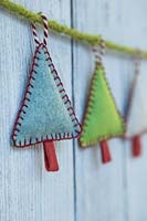 Making stitched felt christmas decorations - miniature christmas trees made from felt and decorative string, hanging against a wooden panel