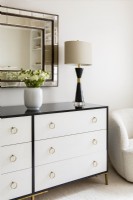 Vase of flowers and lamp on black and white chest of drawers with mirror above.