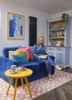 Woman reading on a blue sofa with a crockery cupboard and a pink spotty rug.