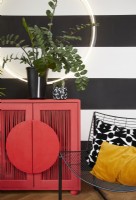 Living room detail showing a modern red cabinet, circle light, contemporary metal chairs and plants. With painted black and white striped walls and a patterned rug.