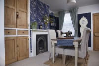 Dining room showing a fireplace, vintage wooden cupboards and blue patterned wallpaper.