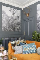 Corner of an orange velvet sofa and green cushions in front of grey door and grey paneled walls with monochrome tropical tree patterned wall papered panels