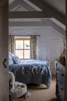 View Into cosy bedroom, bed with blue covers, an old chest of drawers, and whitewashed walls and beams.