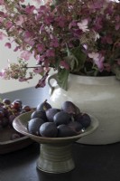 Bowl of fresh figs next to flower arrangement in ceramic pot 