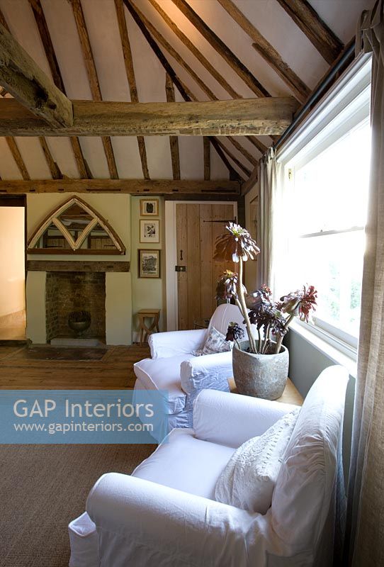 Boonshill Farm, East Sussex. Interior of bedroom with wooden floorboards, exposed beams and linen covered armchairs and mirror made from old window. 