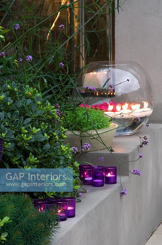 Rendered raised bed beside fireplace with Liriope muscari, candles and a large glass bowl with gerberas in garden at night