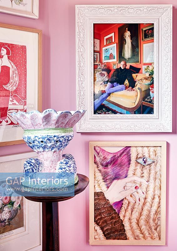Framed photographs and paintings on pink wall with colourful pottery 