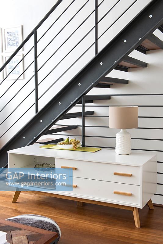 Retro sideboard under open metal staircase with striped wall 