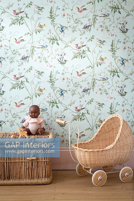 Wicker pram, toy dolly and basket in childrens room with floral wallpaper 