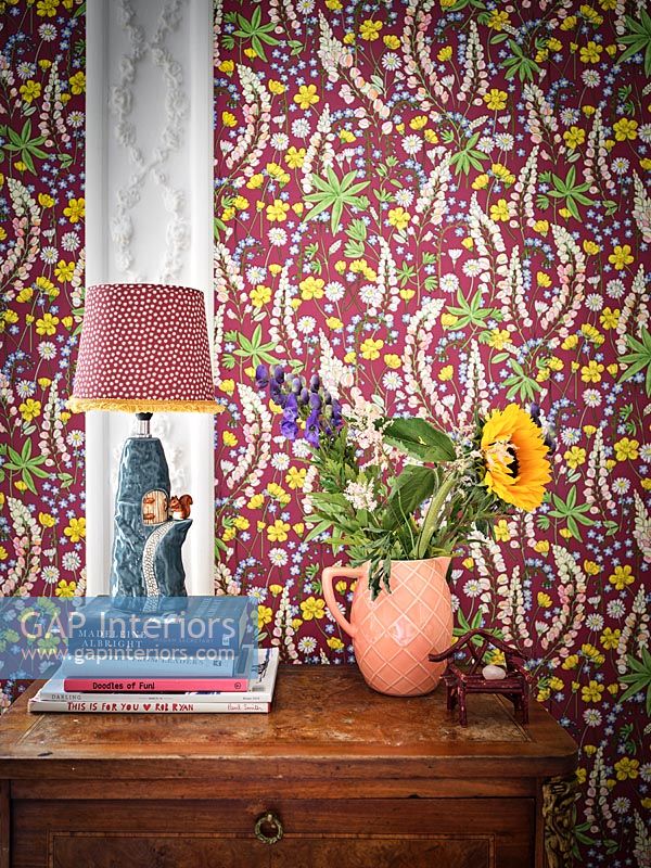 Flowers and lamp on table with colourful floral wallpaper on wall behind