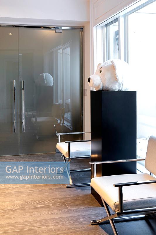 Modern white chairs in hallway next to plinth with large teddy bear head 