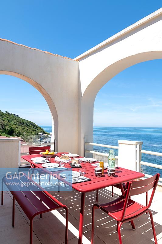 Red dining table and chairs on white terrace with sea views in summer 