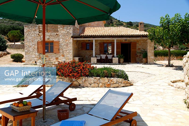 Recliners under shade of parasol on stone paved terrace - country house exterior 