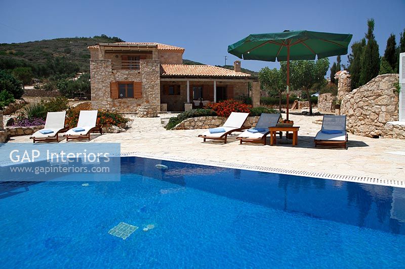 Recliners under shade of parasol next to pool on stone paved terrace 