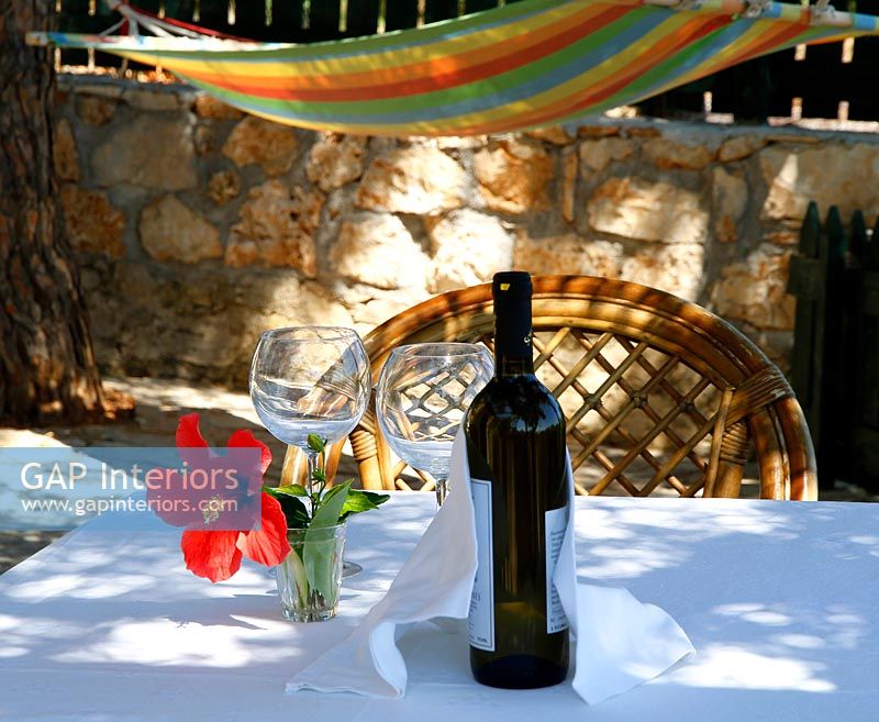Tablecloth and bottle of wine on small garden table shaded by trees with hammock 