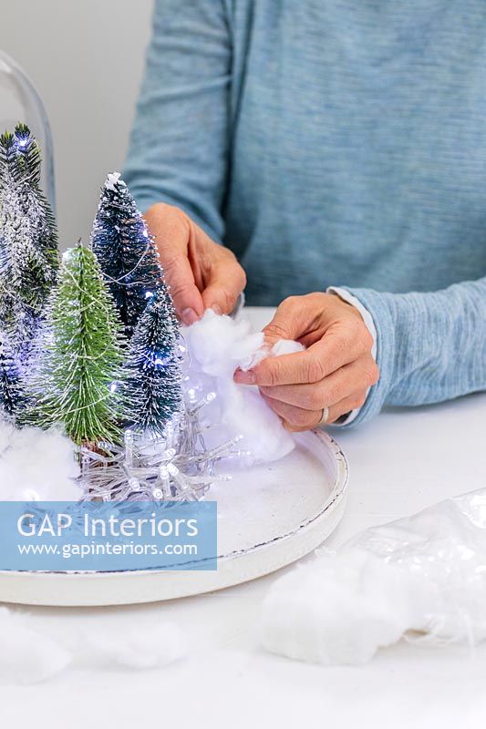 Woman adding cotton wool as snow to tray with miniature christmas trees and fairy light