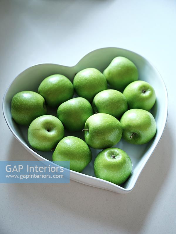 Heart shaped white fruit bowl filled with green apples 