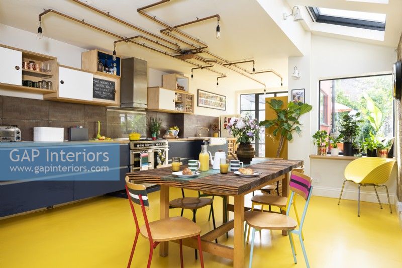 Modern retro kitchen with exposed brick, yellow rubber floor, blue cabinets, plywood, copper pipe lighting and upcycled table and chairs