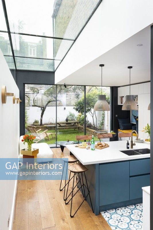 Contemporary modern kitchen in a side return extension with a blue island, wooden floors, light pendants, bar stools and slim frame tall aluminium doors.