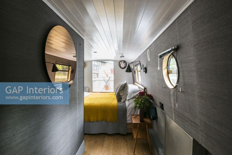 Contemporary bedroom on canal, narrow, river boat with dark grey walls, wooden floors, white painted ceiling, round mirror, porthole window and yellow throw.