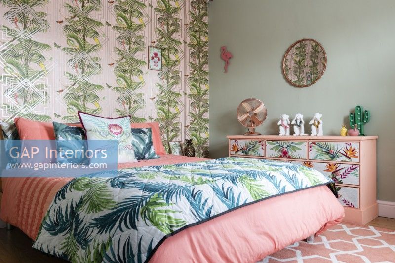 Modern bedroom with palm patterned wallpaper and bedspread and floral painted chest of drawers