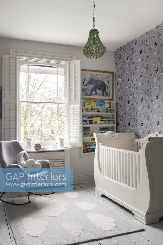 Wooden cot in nursery with paterned wallpaper and rocking chair