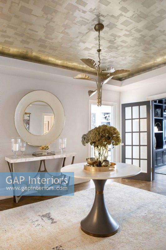 Modern foyer entrance with grey and white tables and gold metal leaf wallpaper on ceiling.