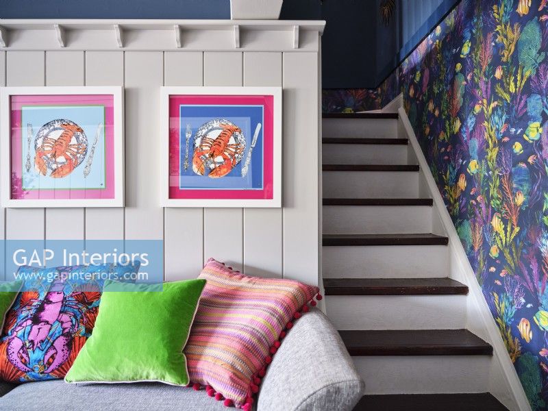 Oceanic artwork on white panelled wall above colourful cushions and next to a small white staircase