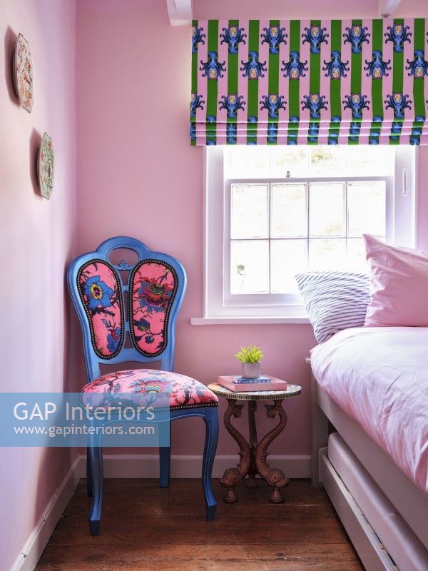 Blue and pink patterned upholstered chair with carved bedside table and colourful blinds 