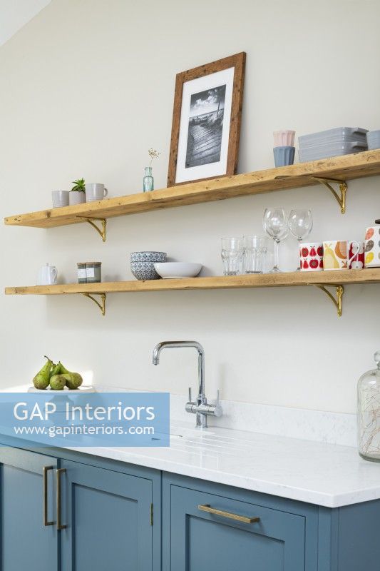 Open wooden shelves in modern classic kitchen with brass brackets, blue units, white countertop and taps.