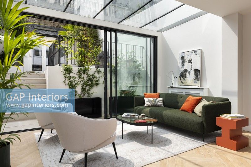 Modern extension conservatory with glass sliding doors, green fabric sofa, orange side table and cushions.