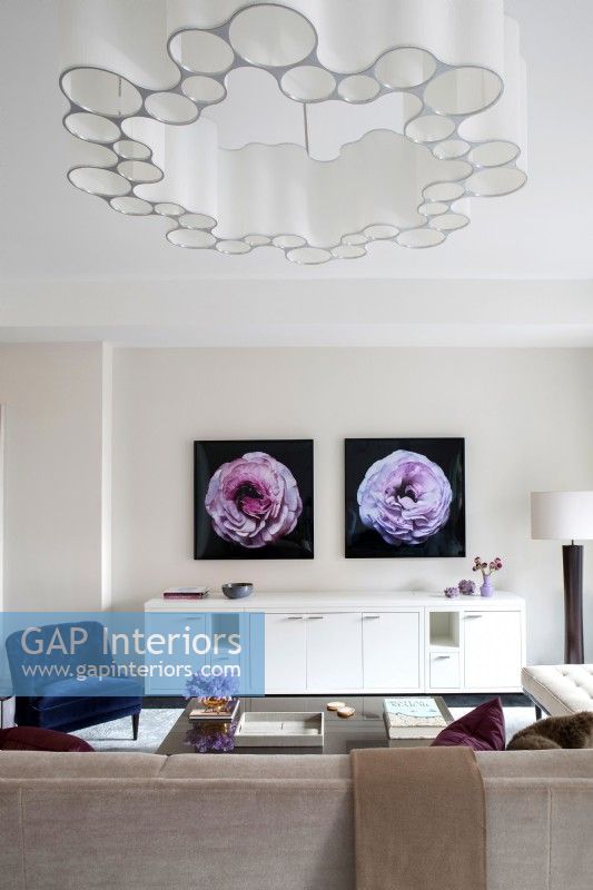 Modern living room with long white credenza decorated with photographs of flowers on wall and unique chandelier.