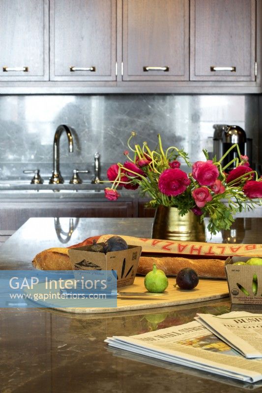Vase of red flowers, food and newspapers displayed on kitchen island.