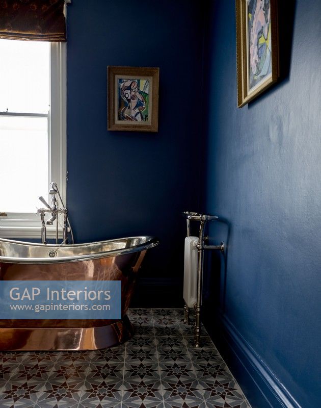 Detail of copper coloured roll top bath in a blue painted bathroom with painting on walls. 