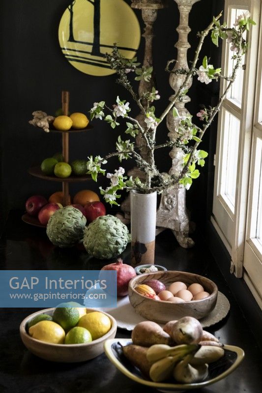 Bowls of fruit next to branches with blossom in vase against black wall