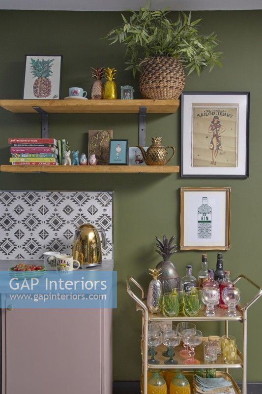 Kitchen detail with a gold kettle, wooden open shelving, green painted walls, patterned tiles and a drinks trolley.