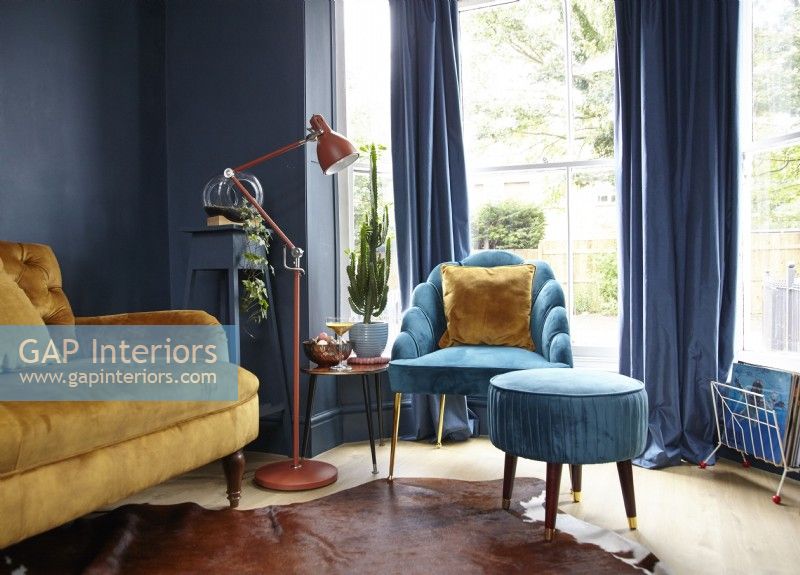 Living room showing a mustard yellow sofa, turquoise armchair with footstool, navy painted wall and a floor lamp. 
