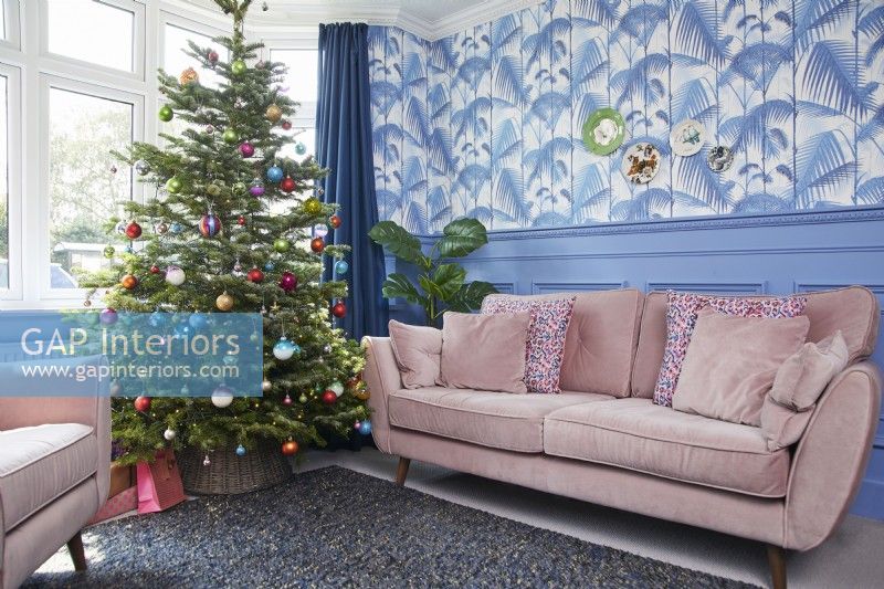 Christmas living room with a decorated tree, pink sofa, jungle print wallpaper and blue panelling.