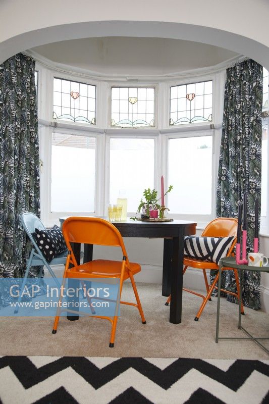 Dining room area with orange and blue chairs in a bay window.