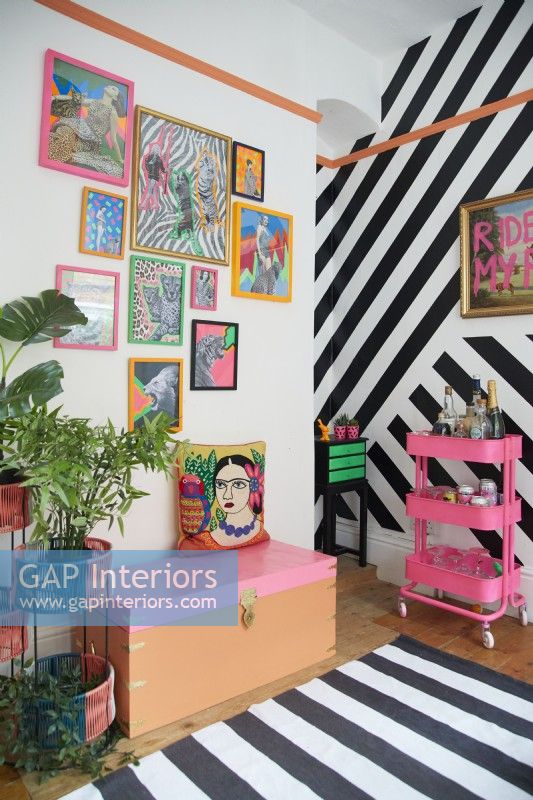 Colourful home office detail with framed artwork, black vinyl striped wall and fluorescent drinks trolley. 