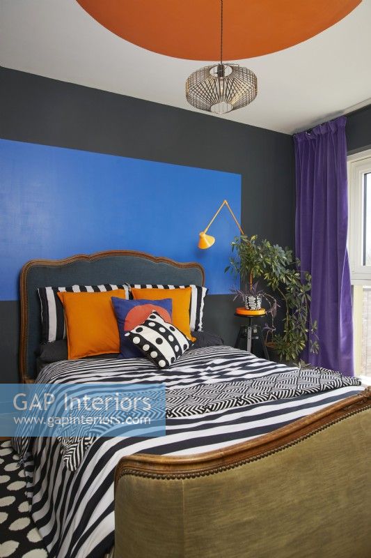 Bold and colourful painted bauhaus shapes in a contemporary bedroom. With an antique headboard, patterned soft furnishing and purple velvet curtains.