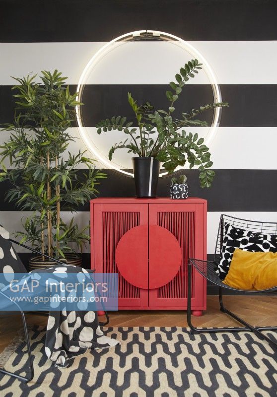 Living room detail showing a modern red cabinet, circle light, contemporary metal chairs and plants. With painted black and white striped walls and a patterned rug.
